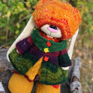 K&ouml;nnte beinhalten: Ein geh&auml;keltes H&auml;schen, das einen bunten Mantel, Hut und Stiefel tr&auml;gt. Das H&auml;schen sitzt auf einem Baumstamm mit gr&uuml;nem und braunem Hintergrund. Der Hut des H&auml;schens ist orange, rot und gelb. Der Mantel ist gr&uuml;n, lila und gelb. Die Stiefel sind gelb. Das H&auml;schen hat eine rosa Nase und schwarze Augen.