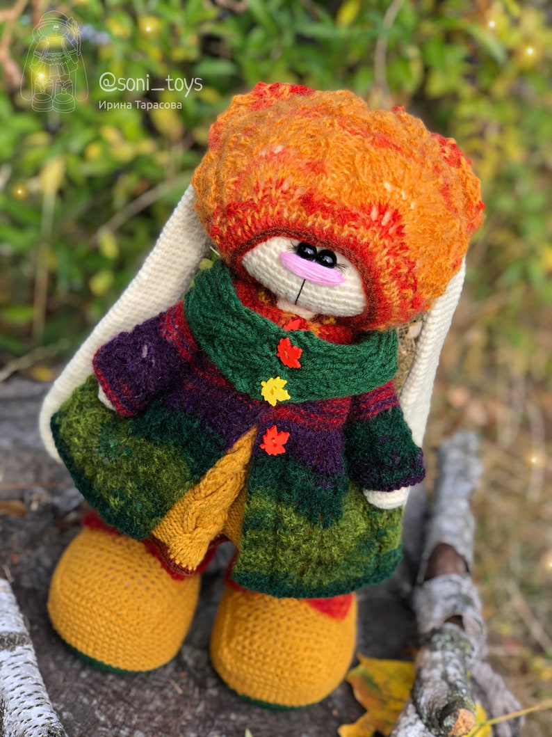 K&ouml;nnte beinhalten: Ein geh&auml;keltes H&auml;schen, das einen bunten Mantel, Hut und Stiefel tr&auml;gt. Das H&auml;schen sitzt auf einem Baumstamm mit gr&uuml;nem und braunem Hintergrund. Der Hut des H&auml;schens ist orange, rot und gelb. Der Mantel ist gr&uuml;n, lila und gelb. Die Stiefel sind gelb. Das H&auml;schen hat eine rosa Nase und schwarze Augen.