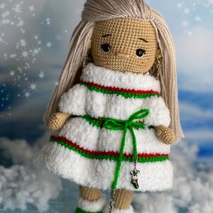Puede incluir: Una mu&ntilde;eca de crochet que lleva un vestido blanco con rayas rojas y verdes, una faja verde y botas verdes. La mu&ntilde;eca tiene el pelo rubio y los ojos marrones. La mu&ntilde;eca sostiene un amuleto plateado en forma de calcet&iacute;n.