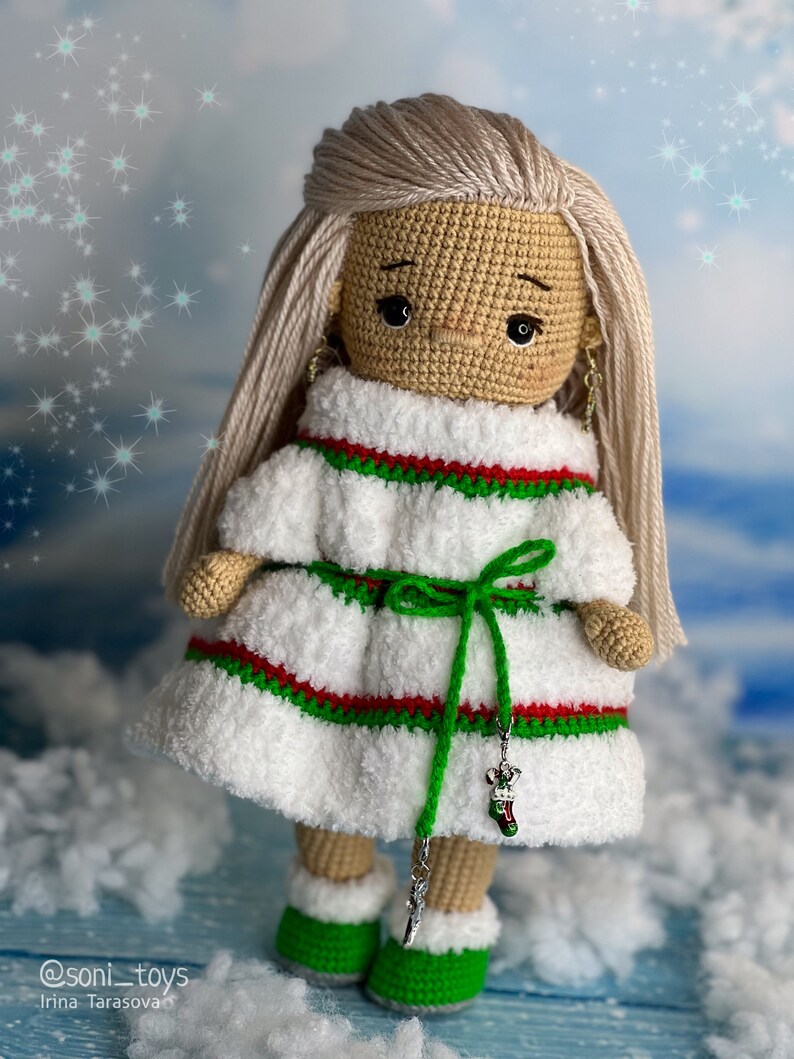 Puede incluir: Una mu&ntilde;eca de crochet que lleva un vestido blanco con rayas rojas y verdes, una faja verde y botas verdes. La mu&ntilde;eca tiene el pelo rubio y los ojos marrones. La mu&ntilde;eca sostiene un amuleto plateado en forma de calcet&iacute;n.