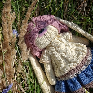 Pode incluir: Um brinquedo de coelho de croch&ecirc; com um chap&eacute;u rosa, um su&eacute;ter branco e uma saia azul e marrom. O coelho est&aacute; deitado em uma cama de grama verde.