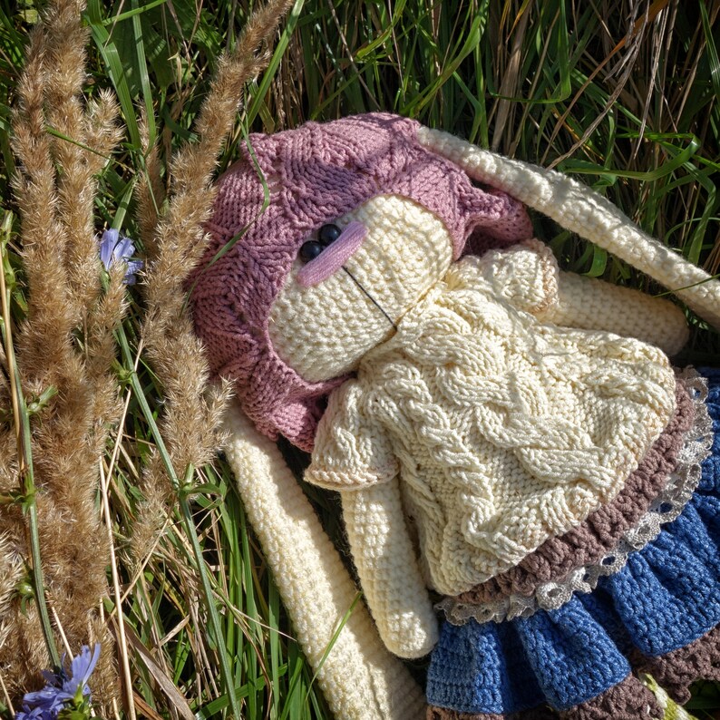 Pode incluir: Um brinquedo de coelho de croch&ecirc; com um chap&eacute;u rosa, um su&eacute;ter branco e uma saia azul e marrom. O coelho est&aacute; deitado em uma cama de grama verde.