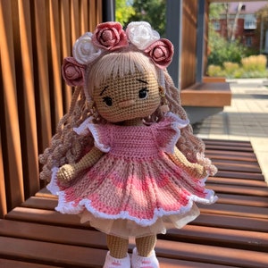 May include: A crocheted doll wearing a pink dress with white lace trim, a flower crown, and white sneakers. The doll has long brown hair and a pink and white flower crown.