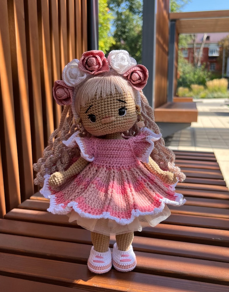 May include: A crocheted doll wearing a pink dress with white lace trim, a flower crown, and white sneakers. The doll has long brown hair and a pink and white flower crown.