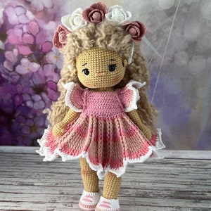 May include: A crocheted doll with blonde curly hair wearing a pink and white dress and a flower crown. The doll is standing on a wooden surface.
