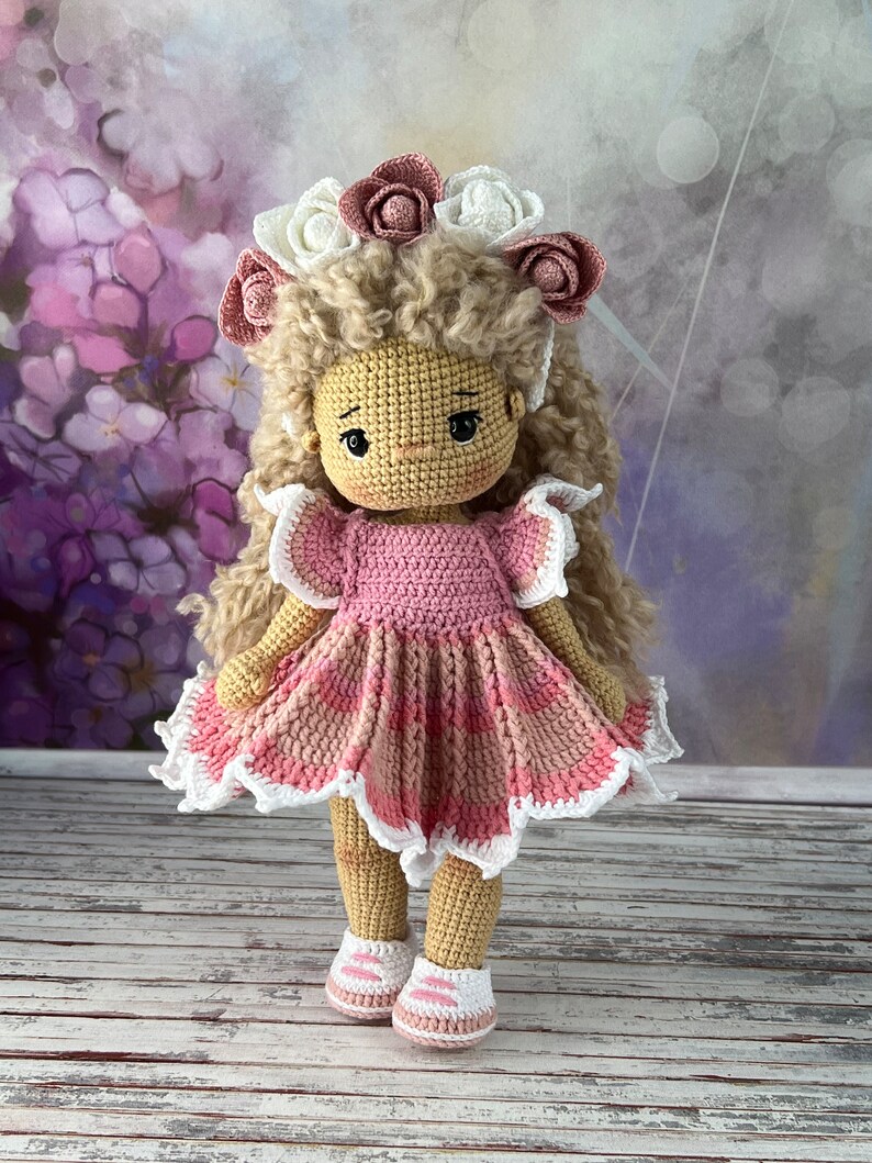 May include: A crocheted doll with blonde curly hair wearing a pink and white dress and a flower crown. The doll is standing on a wooden surface.