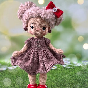 May include: A crocheted doll with brown curly hair, a brown dress, red shoes, and a red bow. The doll is standing on a green grassy surface.