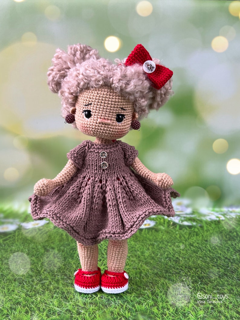 May include: A crocheted doll with brown curly hair, a brown dress, red shoes, and a red bow. The doll is standing on a green grassy surface.