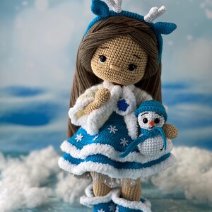 May include: A crocheted doll wearing a blue and white dress with snowflake accents and a white fur trim. The doll has brown hair and is wearing a blue headband with white antlers. The doll is holding a crocheted snowman.