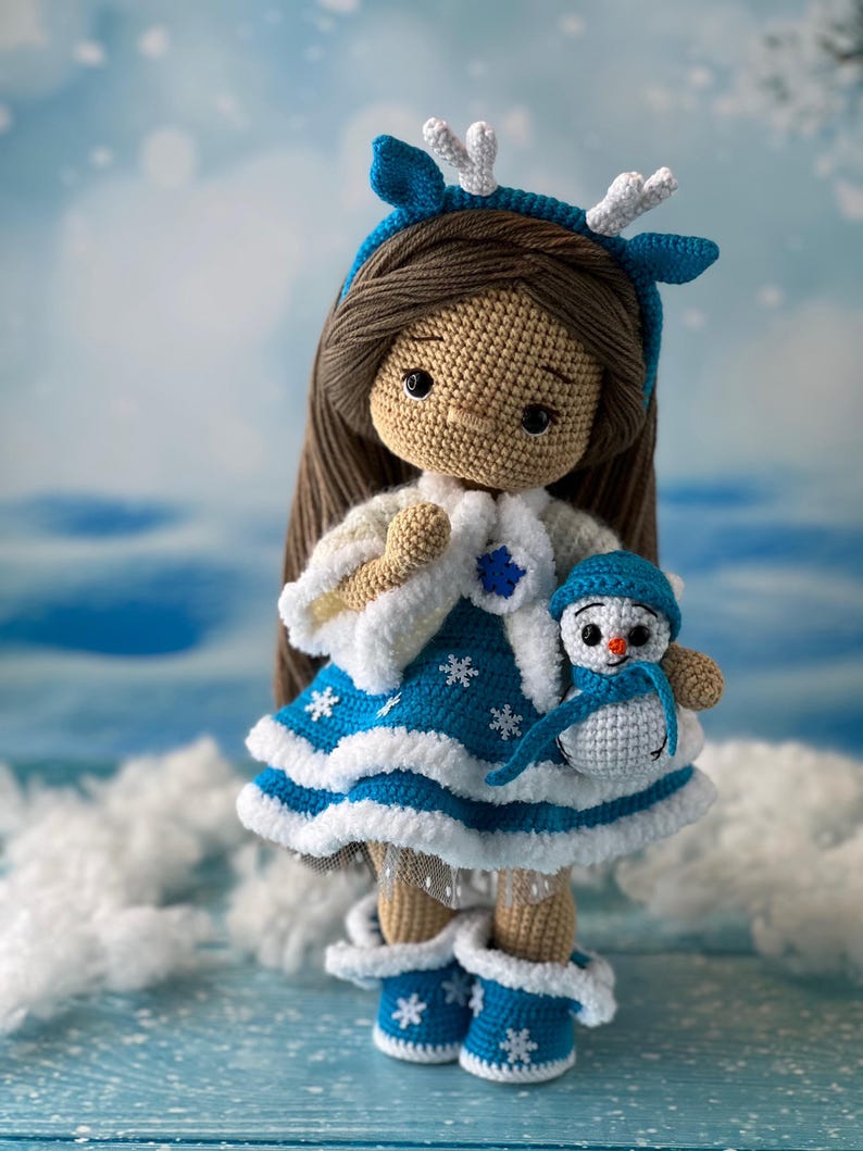 May include: A crocheted doll wearing a blue and white dress with snowflake accents and a white fur trim. The doll has brown hair and is wearing a blue headband with white antlers. The doll is holding a crocheted snowman.