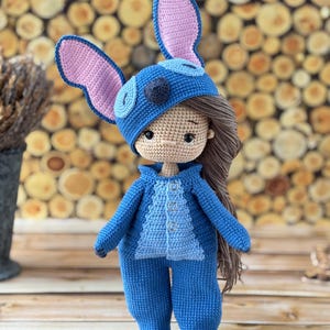 Pu&ograve; includere: Bambola all'uncinetto fatta a mano, vestita con una tuta e un cappello blu con grandi orecchie foderate di rosa. La bambola ha i capelli castani ed &egrave; appoggiata su una superficie di legno. L'outfit della bambola ha un davanti abbottonato.