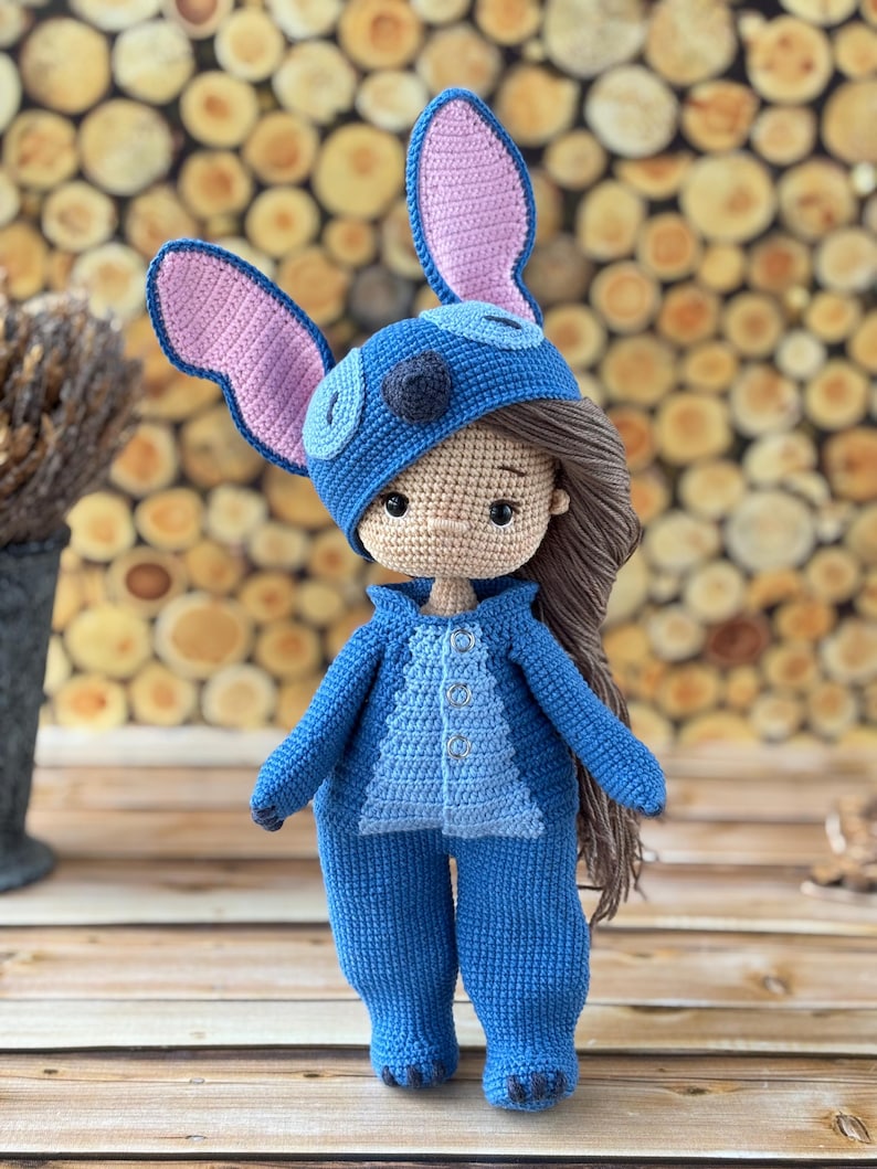 Pu&ograve; includere: Bambola all'uncinetto fatta a mano, vestita con una tuta e un cappello blu con grandi orecchie foderate di rosa. La bambola ha i capelli castani ed &egrave; appoggiata su una superficie di legno. L'outfit della bambola ha un davanti abbottonato.