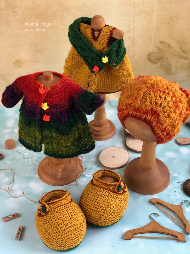 K&ouml;nnte beinhalten: Ein Set gestrickter Puppenkleidung in Herbstfarben, bestehend aus einem Kleid, einem Cardigan, einer M&uuml;tze und Stiefeletten. Die Kleidung wird auf Holzpuppen und Kleiderb&uuml;geln pr&auml;sentiert. @soni_toys Irina Tarasova