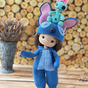 Pu&ograve; includere: Bambola all'uncinetto fatta a mano, vestita con tuta e cappello blu con grandi orecchie. Una bambola turchese pi&ugrave; piccola con un fiocco rosa &egrave; in cima al cappello. La bambola ha i capelli castani e un incarnato neutro. Sfondo in legno.