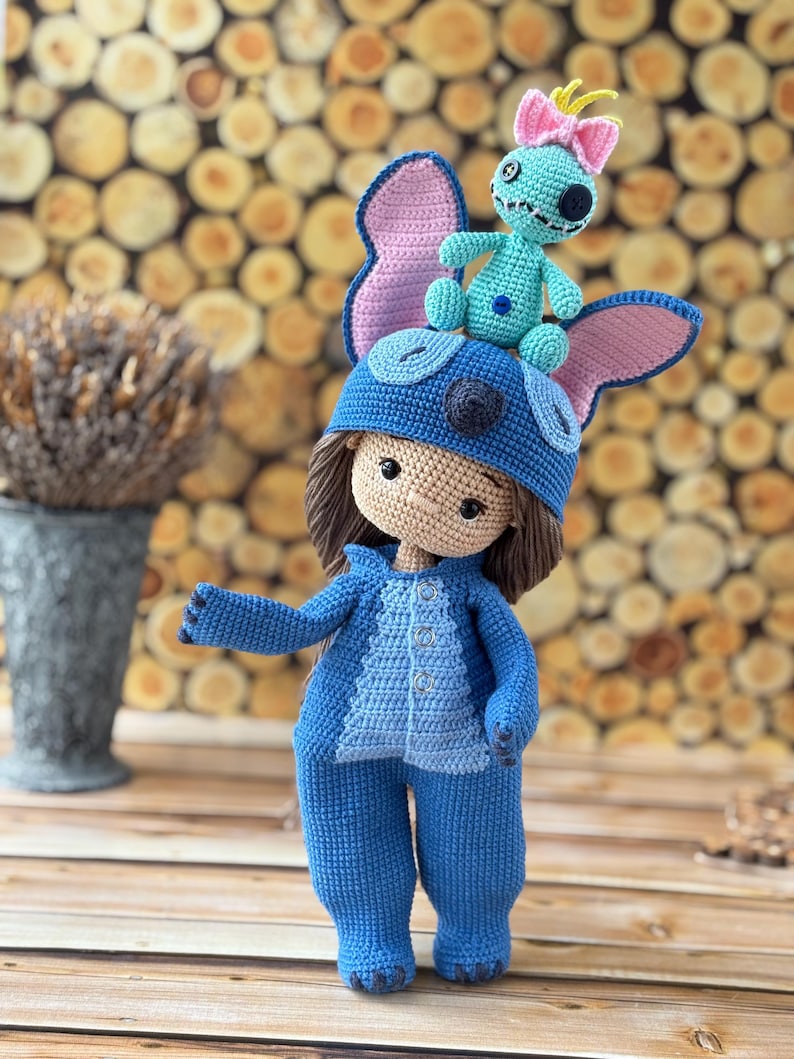 Pu&ograve; includere: Bambola all'uncinetto fatta a mano, vestita con tuta e cappello blu con grandi orecchie. Una bambola turchese pi&ugrave; piccola con un fiocco rosa &egrave; in cima al cappello. La bambola ha i capelli castani e un incarnato neutro. Sfondo in legno.