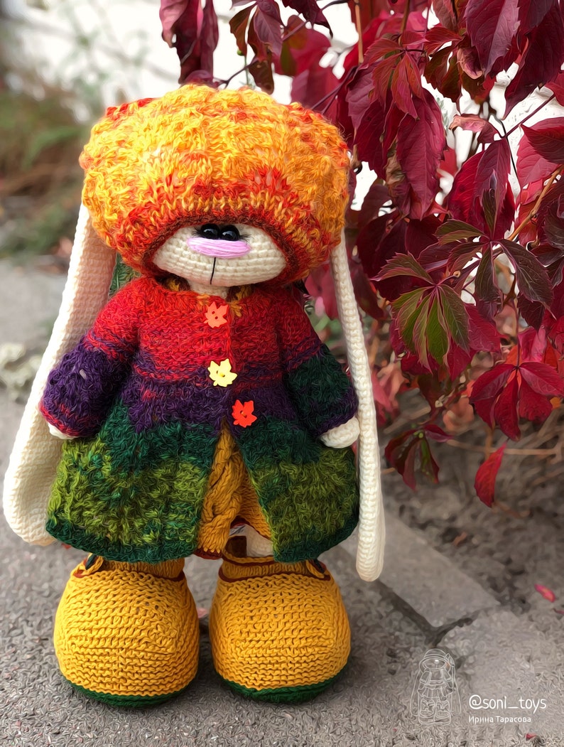 K&ouml;nnte beinhalten: Ein geh&auml;keltes H&auml;schen, das einen mehrfarbigen Pullover, eine M&uuml;tze und gelbe Schuhe tr&auml;gt. Das H&auml;schen steht vor einem Busch mit roten Bl&auml;ttern. @soni_toys Irina Tarasova