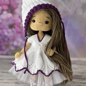 May include: A crocheted doll with long brown hair, wearing a white dress with a purple trim, a white hat with a purple trim, and white shoes with purple trim.