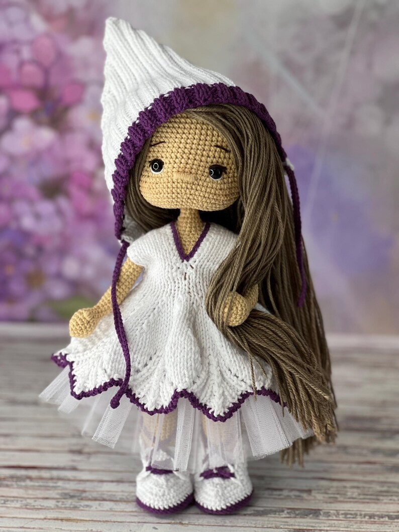 May include: A crocheted doll with long brown hair, wearing a white dress with a purple trim, a white hat with a purple trim, and white shoes with purple trim.