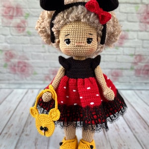 May include: A crocheted doll with brown curly hair, wearing a red and black polka dot dress with black lace trim, black shoes, and a yellow crocheted purse. The doll is wearing a black headband with red bow and black mouse ears.