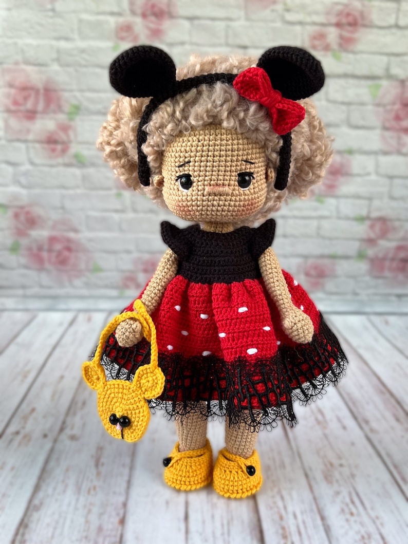 May include: A crocheted doll with brown curly hair, wearing a red and black polka dot dress with black lace trim, black shoes, and a yellow crocheted purse. The doll is wearing a black headband with red bow and black mouse ears.