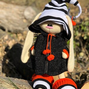 Puede incluir: Un conejito de crochet con un sombrero a rayas blanco y negro, una chaqueta negra con pompones naranjas y botines a rayas naranja y negro.