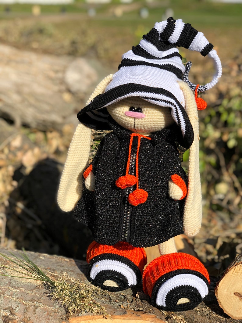 Puede incluir: Un conejito de crochet con un sombrero a rayas blanco y negro, una chaqueta negra con pompones naranjas y botines a rayas naranja y negro.