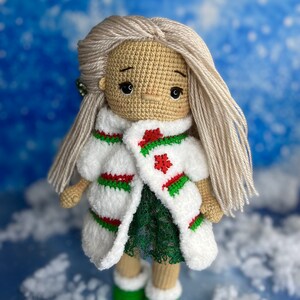 Puede incluir: Una mu&ntilde;eca de crochet con el pelo largo y rubio que lleva un abrigo blanco y verde con un dise&ntilde;o de copo de nieve rojo. La mu&ntilde;eca lleva un vestido verde y botas verdes.