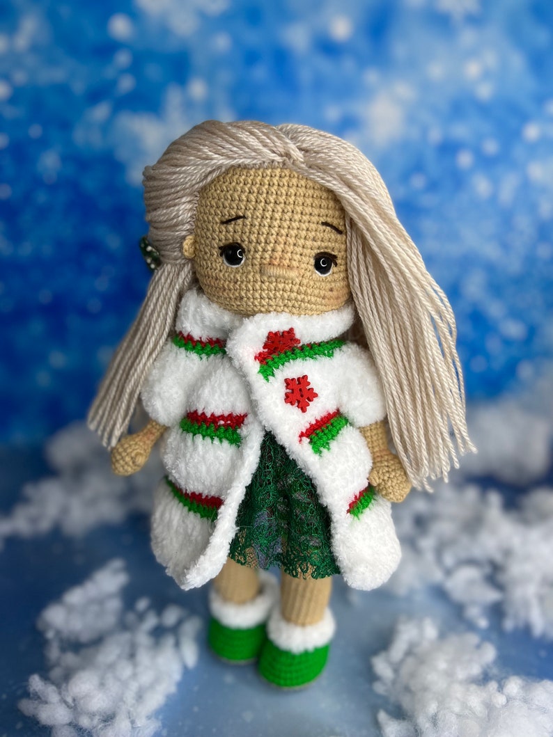 Puede incluir: Una mu&ntilde;eca de crochet con el pelo largo y rubio que lleva un abrigo blanco y verde con un dise&ntilde;o de copo de nieve rojo. La mu&ntilde;eca lleva un vestido verde y botas verdes.
