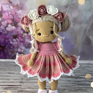 May include: A crocheted doll with blonde hair wearing a pink and white dress and a flower crown. The doll is wearing pink and white shoes and is standing on a wooden surface.