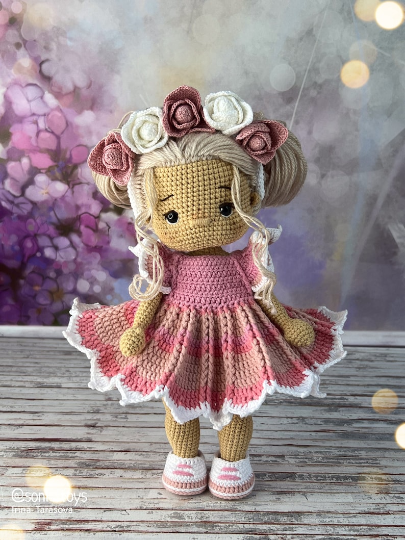 May include: A crocheted doll with blonde hair wearing a pink and white dress and a flower crown. The doll is wearing pink and white shoes and is standing on a wooden surface.