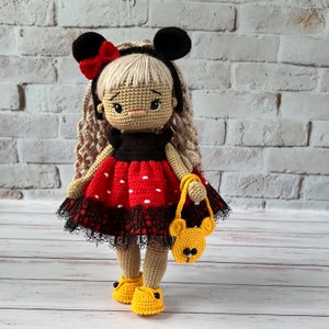 May include: A crocheted doll with blonde hair, wearing a red and black polka dot dress with a black lace trim, and black mouse ears with a red bow. The doll is holding a yellow crocheted purse with a bear face.