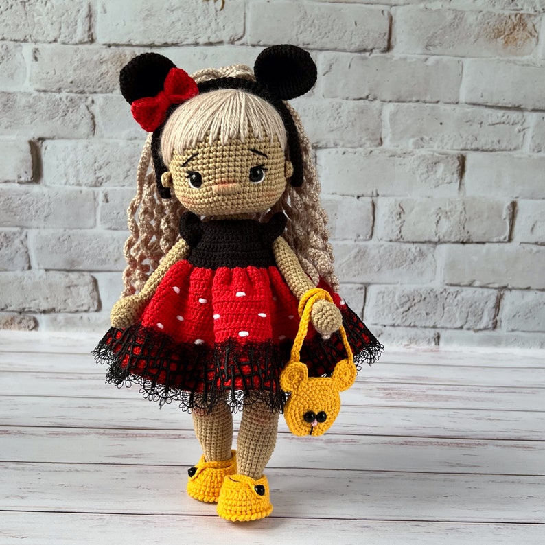 May include: A crocheted doll with blonde hair, wearing a red and black polka dot dress with a black lace trim, and black mouse ears with a red bow. The doll is holding a yellow crocheted purse with a bear face.