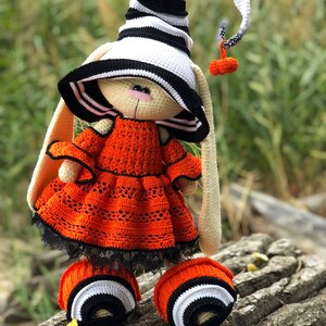 Puede incluir: Un conejito de crochet de Halloween con un sombrero de bruja a rayas blancas y negras, un vestido naranja con ribete de encaje negro y botines a rayas blancas y negras.