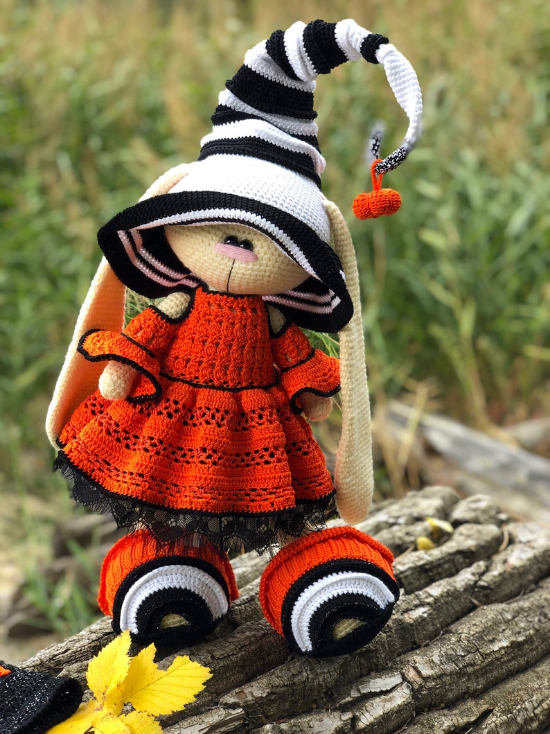 Puede incluir: Un conejito de crochet de Halloween con un sombrero de bruja a rayas blancas y negras, un vestido naranja con ribete de encaje negro y botines a rayas blancas y negras.