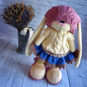 Pode incluir: Um brinquedo de coelho de croch&ecirc; vestindo um chap&eacute;u rosa, um su&eacute;ter amarelo e uma saia azul com acabamento de renda marrom. O coelho tem orelhas longas e est&aacute; em p&eacute; sobre uma superf&iacute;cie de madeira branca.