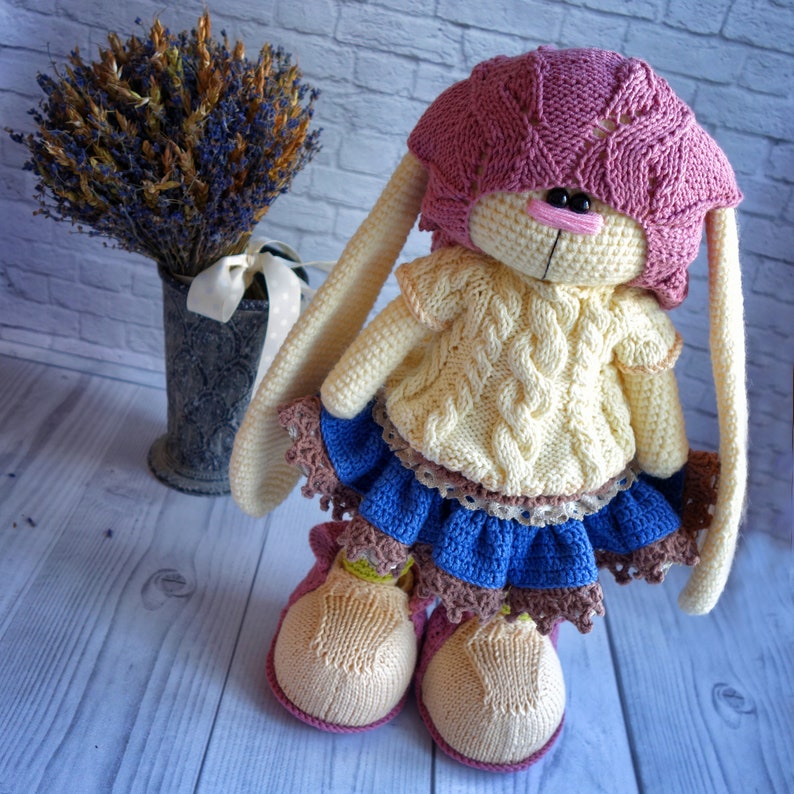 Pode incluir: Um brinquedo de coelho de croch&ecirc; vestindo um chap&eacute;u rosa, um su&eacute;ter amarelo e uma saia azul com acabamento de renda marrom. O coelho tem orelhas longas e est&aacute; em p&eacute; sobre uma superf&iacute;cie de madeira branca.