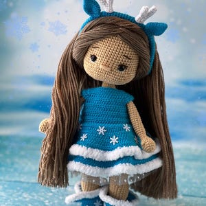 May include: A crocheted doll wearing a blue dress with white snowflakes, a white headband with antlers, and blue boots with white snowflakes. The doll has brown hair and a beige face.