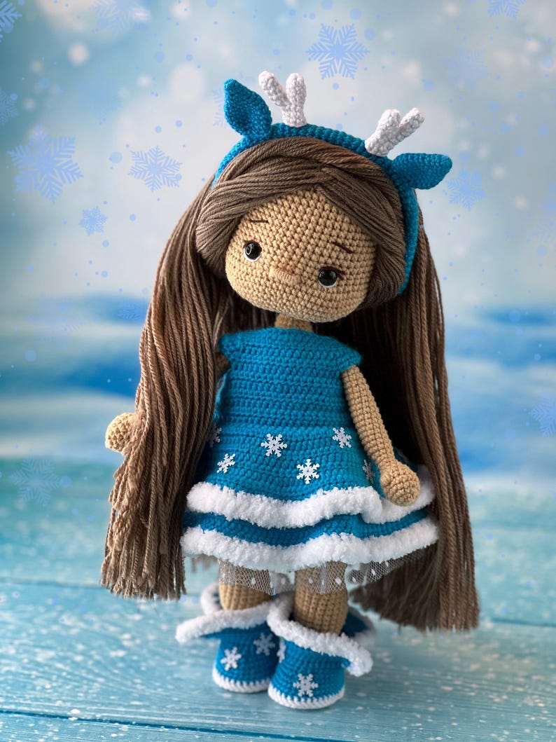 May include: A crocheted doll wearing a blue dress with white snowflakes, a white headband with antlers, and blue boots with white snowflakes. The doll has brown hair and a beige face.