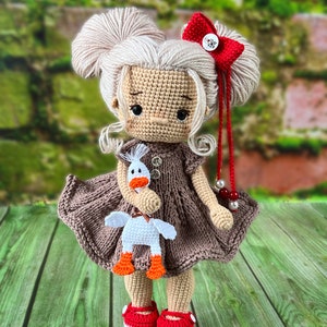 May include: A crocheted doll with blonde hair, a brown dress, and red shoes. The doll is holding a white crocheted duck with orange feet.
