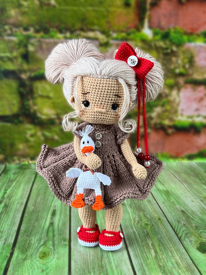 May include: A crocheted doll with blonde hair, a brown dress, and red shoes. The doll is holding a white crocheted duck with orange feet.