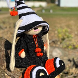 Puede incluir: Un conejito de crochet de Halloween con un sombrero a rayas blanco y negro con un pomp&oacute;n naranja. El conejito lleva un atuendo negro y naranja con pompones naranjas en la parte delantera.