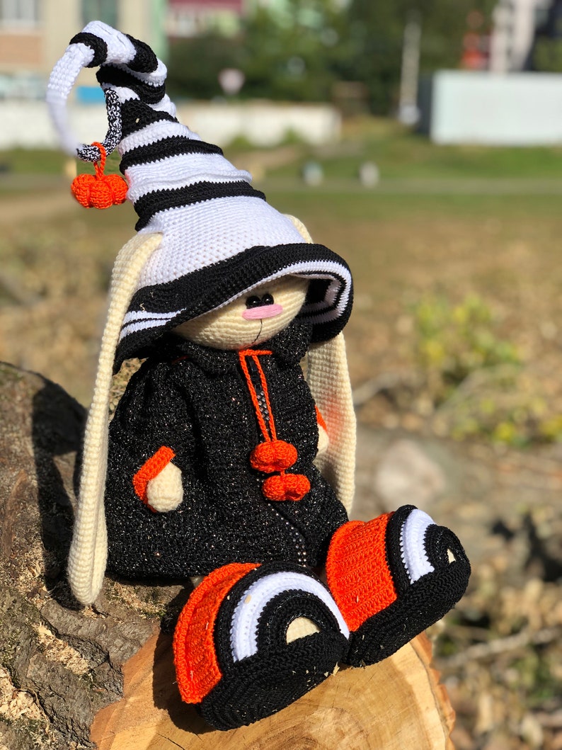 Puede incluir: Un conejito de crochet de Halloween con un sombrero a rayas blanco y negro con un pomp&oacute;n naranja. El conejito lleva un atuendo negro y naranja con pompones naranjas en la parte delantera.