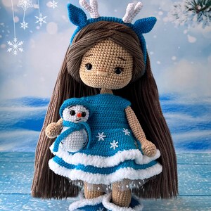 May include: A crocheted doll wearing a blue dress with white trim and a reindeer headband. The doll is holding a small crocheted snowman. The background is a blue and white winter scene with snowflakes.