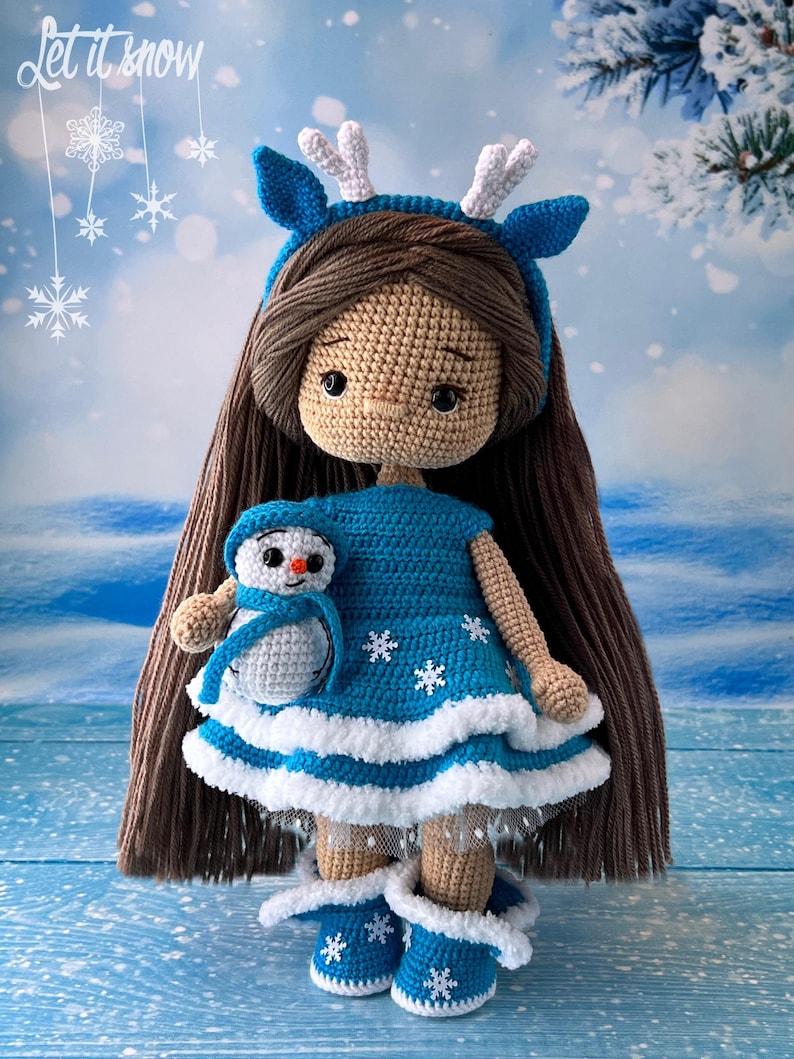 May include: A crocheted doll wearing a blue dress with white trim and a reindeer headband. The doll is holding a small crocheted snowman. The background is a blue and white winter scene with snowflakes.