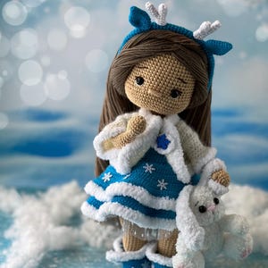 May include: A crocheted doll wearing a blue and white dress with a white fur trim, a blue and white headband with white antlers, and blue and white boots. The doll is holding a white crocheted bunny.