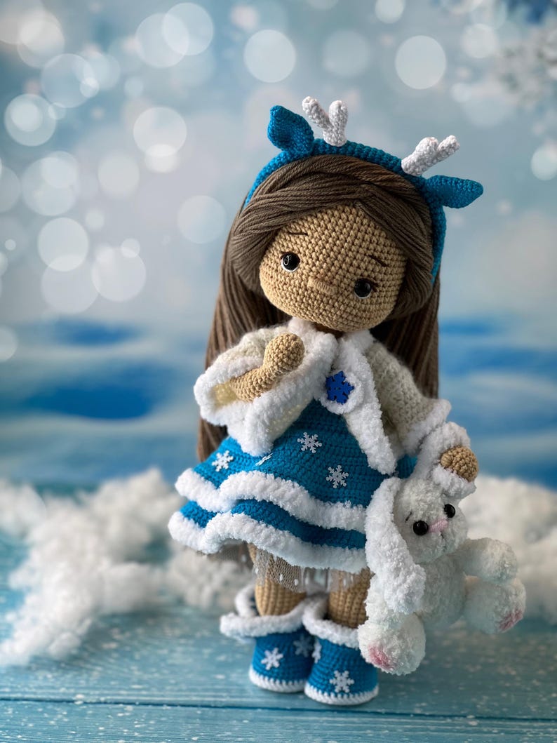 May include: A crocheted doll wearing a blue and white dress with a white fur trim, a blue and white headband with white antlers, and blue and white boots. The doll is holding a white crocheted bunny.