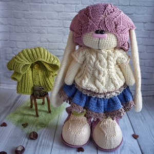 Pode incluir: Um brinquedo de coelho de croch&ecirc; vestindo um chap&eacute;u rosa, um su&eacute;ter creme, uma saia azul e sapatos creme. O coelho tem orelhas brancas longas e um nariz rosa.