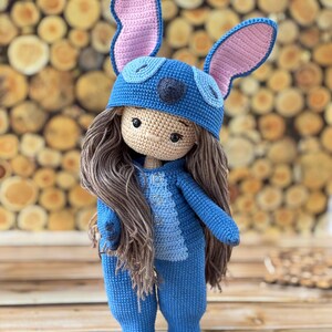 Pu&ograve; includere: Bambola all'uncinetto fatta a mano con una tuta blu e un cappello abbinato con grandi orecchie foderate di rosa. La bambola ha lunghi capelli castani e un incarnato neutro. L'outfit della bambola presenta dettagli di bottoni e un pannello blu chiaro sul petto.