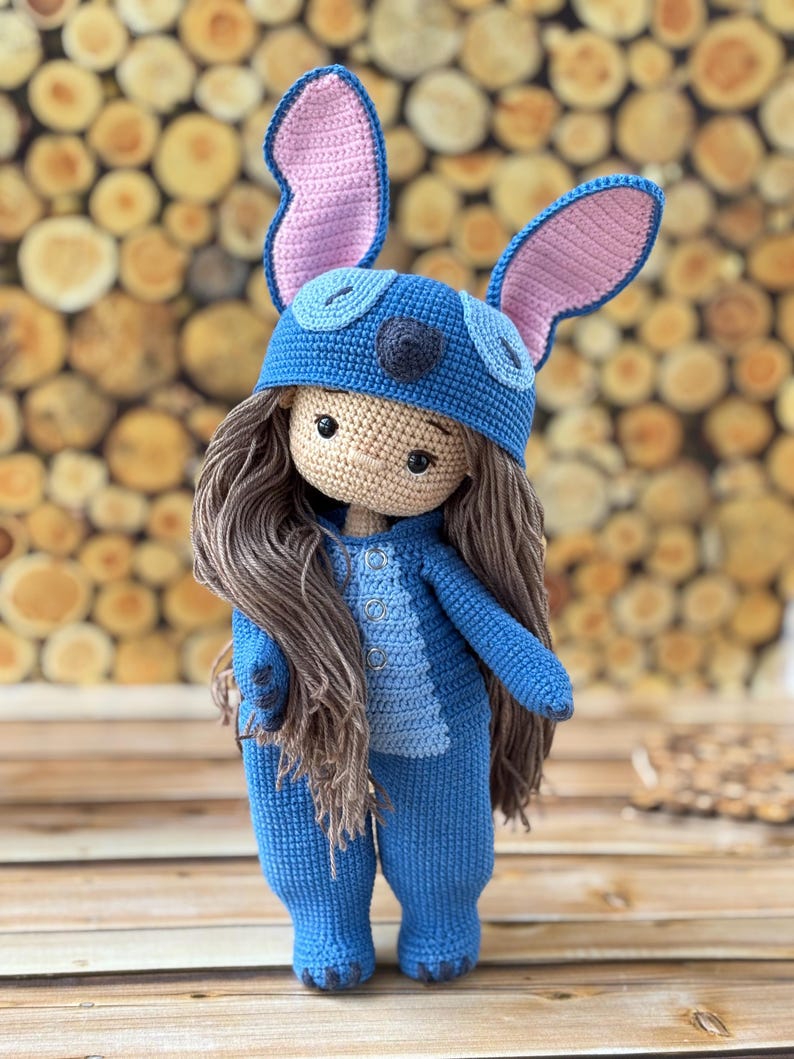Pu&ograve; includere: Bambola all'uncinetto fatta a mano con una tuta blu e un cappello abbinato con grandi orecchie foderate di rosa. La bambola ha lunghi capelli castani e un incarnato neutro. L'outfit della bambola presenta dettagli di bottoni e un pannello blu chiaro sul petto.
