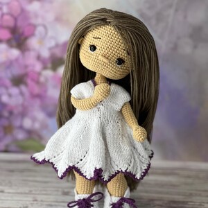May include: A crocheted doll with brown hair, wearing a white dress with purple trim and white shoes with purple laces.